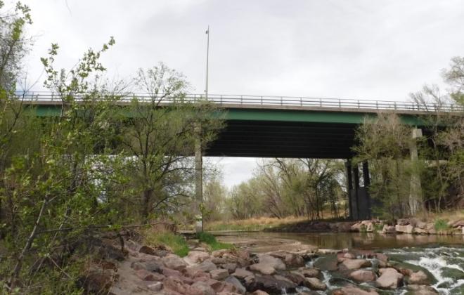 Photo of looking up at a bridge over a rocky creek with two concrete piers and green girders with metal rails and streetlights. 