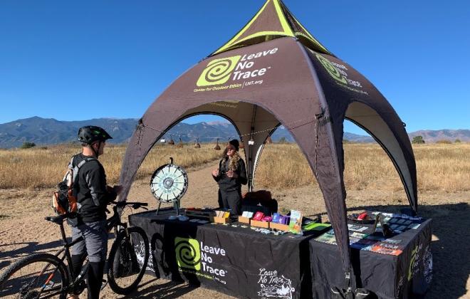 a person on a bike stops and talks to a Leave No Trace representative at a booth in an open space.